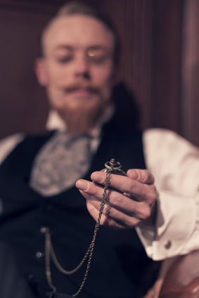 Vintage 1900 fashion man with beard and glasses. Sitting in old wooden reading room. Looking at pocket watch.